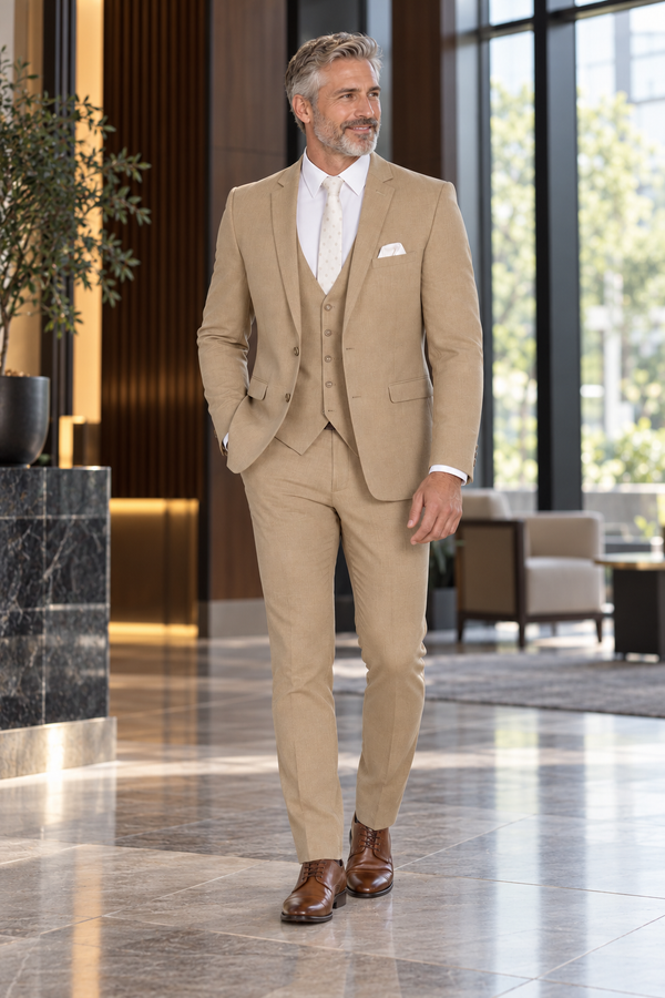 "The Metro" Men's Slim Fit Camel 3 Piece Suit — model photograph in a modern hotel lobby with dark wood vertical panels and tall glass windows, neutral daylight, no crowd